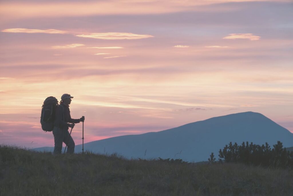 Eine Frau, die einen Rucksack trägt, wandert auf einer Anhöhe mit Blick auf ein Bergpanorama in Abendstimmung.