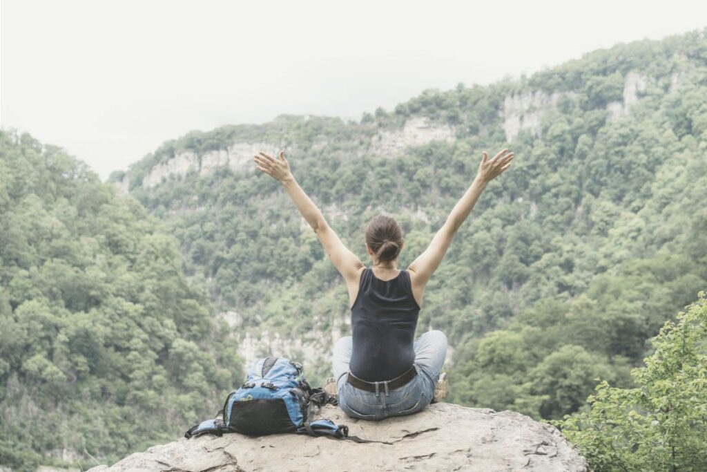 blogartikel 9 2 Eine Frau sitzt auf einem Felsvorsprung mit Bergblick. Ihr Rucksack steht neben ihr, und sie hebt beide Arme in die Luft als Symbol für die Entlastung, nicht für alles verantwortlich zu sein.